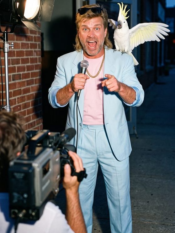 Manny in a baby-blue suit yelling into a microphone at a cameraman, Mr. Feathers flapping on his shoulder